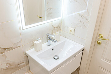 Bright bathroom with a white sink, vanity, and marble-patterned tiles. A lighted mirror and golden door hardware add a touch of modern elegance