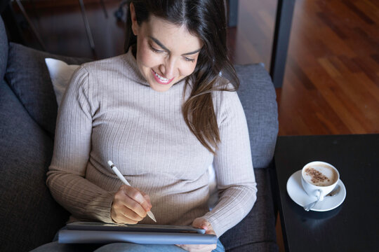 Woman smiling while creating digital art with a stylus and tablet on a sofa