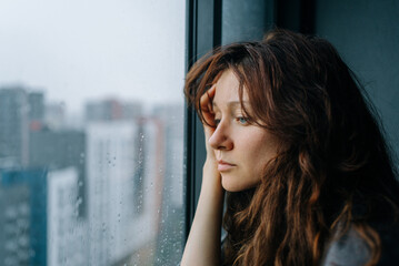Young woman feeling sad and pensive, experiencing autumn depression while looking out a rainy city...