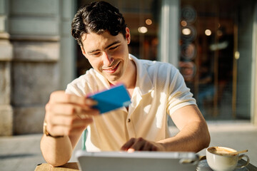 Portrait of a smiling young man guy with a coffee cup in a coffee shop or a restaurant in the city, shopping online using a credit card and tablet online, or entrepreneur businessman working