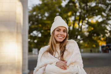 attractive smiling young blond woman walking in winter park having fun in warm white knitted sweater, cape and hat, cold season