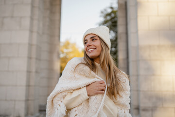 attractive smiling young blond woman walking in winter park having fun in warm white knitted sweater, cape and hat, cold season