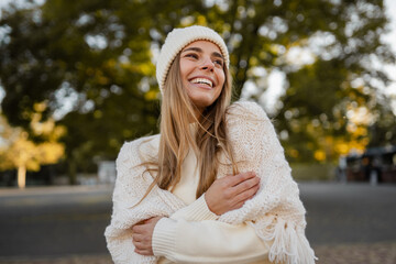 attractive smiling young blond woman walking in winter park having fun in warm white knitted sweater, cape and hat, cold season