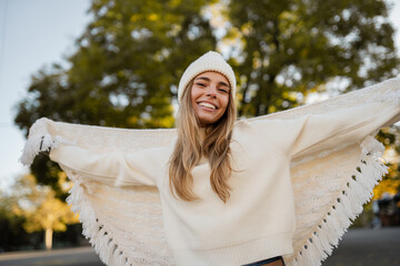 attractive smiling young blond woman walking in winter park having fun in warm white knitted sweater, cape and hat, cold season