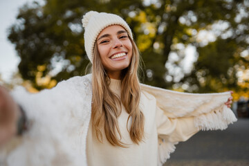 attractive smiling young blond woman walking in winter park having fun in warm white knitted sweater, cape and hat, cold season