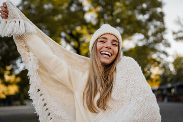 attractive smiling young blond woman walking in winter park having fun in warm white knitted sweater, cape and hat, cold season