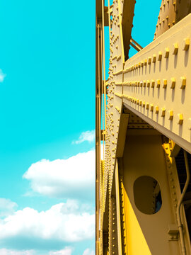 Looking Up At The Side of a Yellow Steel Bridge Into a Turquoise Sky with Fluffy Clouds