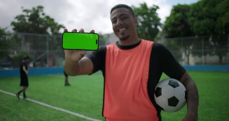 Smiling Latin Hispanic man of African descent showing smartphone with green screen to camera while holding soccer ball on field, technology mockup for mobile app or social network, active leisure - Powered by Adobe