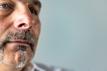 Detailed close-up of a middle-aged man’s lower face, showing the texture of his skin, mustache, and goatee with a neutral blue backdrop. Sad, lonely look.