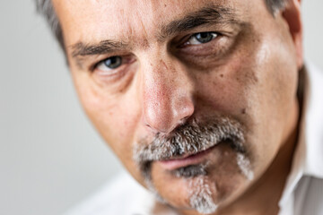 Detailed close-up of a mature man’s face with gray mustache and goatee. The intense gaze and natural lighting emphasize skin detail, facial expression, and realism against a minimal background.
