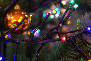 Colorful fairy lights are wrapped around green Christmas tree branches. The lights create a festive atmosphere. A blurred shiny orange bauble hangs in the background