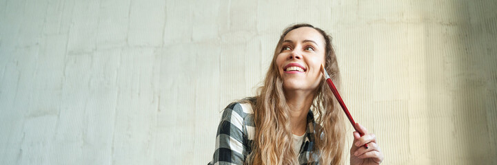 Young caucasian female artist smiling with paintbrush in hand wearing checkered shirt against...