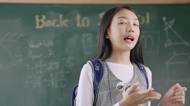 Back to school concept. Portrait of happy Asian child girl student in uniform with a school bag and backpack standing in front of a blackboard with a myself speech and presentation in the classroom. 