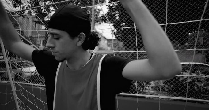 Hispanic soccer player leans against goal net in quiet reflection after game, lost in thought about life and purpose, expressing solitude and introspection on empty urban field, monochrome
