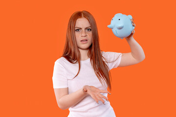 Young woman expressing frustration while holding a blue piggy bank against a bright orange background