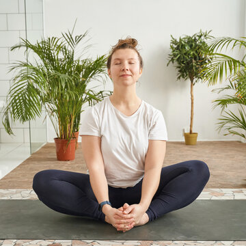 Caucasian young adult female meditating in yoga pose at indoor studio with green plants.
