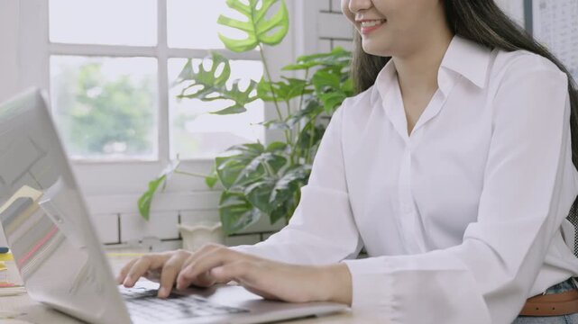 Happy young Asian female teacher sitting smiling and using laptop computer while typing on keyboard at her desk in classroom. Close up business woman working in office. Girl student in homeschool. 