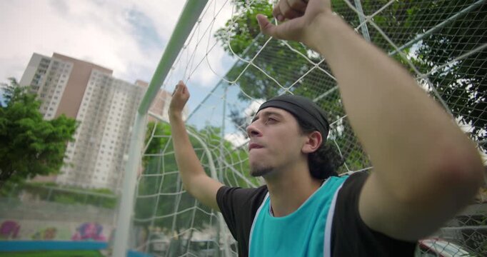 Hispanic soccer player leans on goal net with eyes down, deep in thought after game, expressing quiet contemplation and emotional reflection on city field