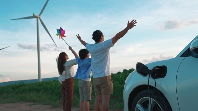 Concept of progressive happy family enjoying their time at wind farm with electric vehicle. Electric vehicle driven by clean renewable energy from wind turbine generator for charging station.