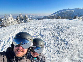Two individuals wearing helmets and ski gear are posing for a selfie on a snowy mountain slope, surrounded by trees and a clear blue sky, capturing a joyful winter adventure