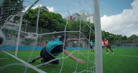 Wide angle view of soccer ball entering goal as Hispanic goalkeeper misses save during community match, showing full field, teammates, and joyful atmosphere of casual play
