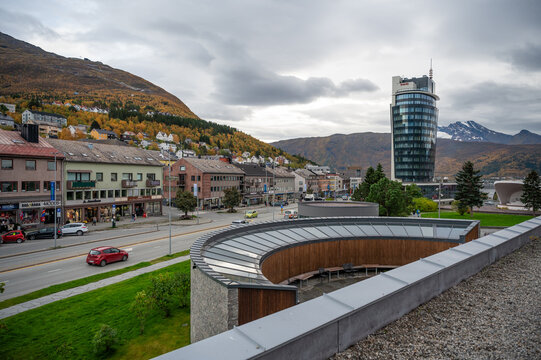 Narvik, Norway, modern building with a curved glass facade stands in a town surrounded by hills and autumn foliage, with a busy street and parked cars