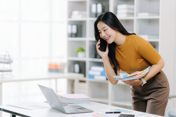 Efficiency and Dialogue: An Asian professional engages in a phone conversation at their work station, multitasking and problem-solving with a radiant smile.