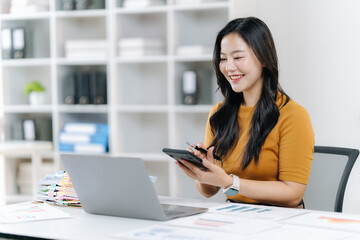 Asian Woman in Home Office: An Asian woman, radiant with a smile, calculates on a calculator while looking at a laptop computer, embodying focused work in a comfortable home environment.