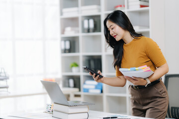 Focused Woman at Workspace: A focused professional woman stands at her modern workstation, intently using her smartphone while managing essential documents.