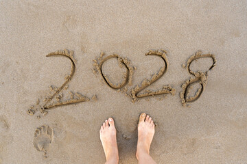 2028 numbers written on sandy beach. Feet top view Sea background.