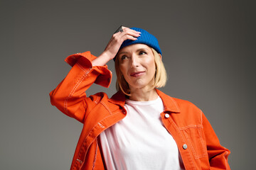 Stylish blonde woman poses in trendy orange jacket and blue beanie in a studio setting