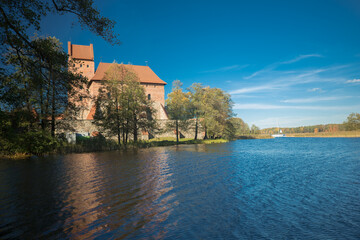 A horizontal shot taken from the water shows Trakai Castle against a clear blue sky. Built of red brick and stone, the castle rises above Lake Galve, surrounded by a fortified wall.

