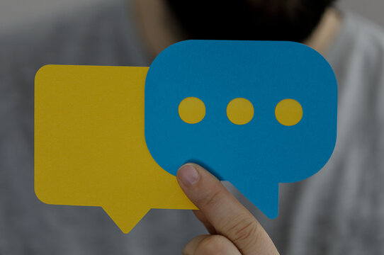 A person holds up two dialog bubbles with dots, a blue one and a yellow one, representing communication, conversation and messaging concepts.
