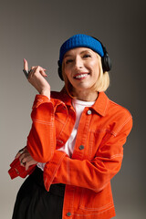 Stylish blonde woman poses in trendy beanie and orange jacket in modern studio setting