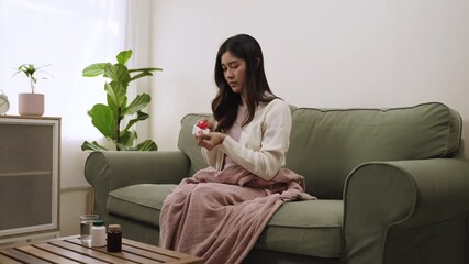 A young Asian woman is lying on the sofa with a blanket because she is sick and has a cough from the influenza while taking medicine to relieve symptoms in the living room of her home. - Powered by Adobe