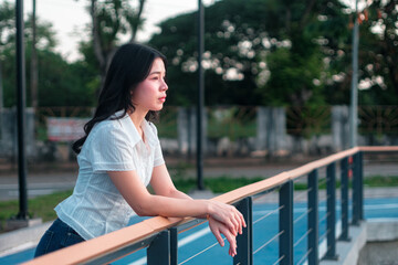 Thoughtful young woman gazes into the distance while leaning on a wooden railing overlooking a serene outdoor landscape during sunset