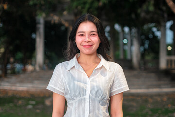 Young woman smiling in a casual white shirt standing outdoors in a park setting with blurred background filled with trees and soft lighting
