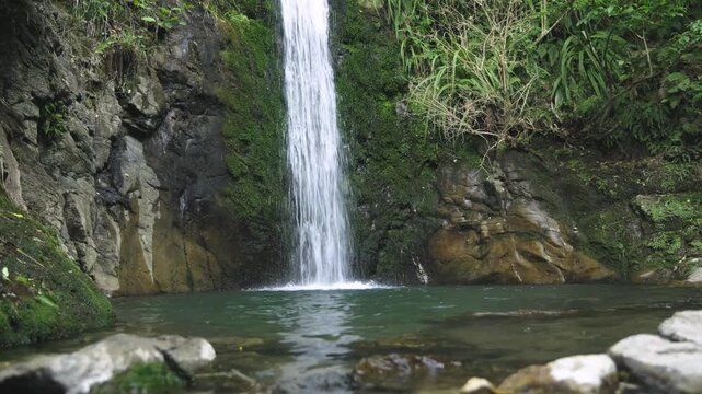The bottom part of a natural waterfall, sloping down a mossy rock, into a small water pool in a lush environment. some out of focus rocks apparent at the foreground. a 4K video clip.