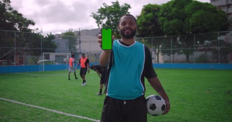 Smiling Hispanic man holding smartphone with green screen toward camera on soccer field, showing mobile app display while holding ball, representing technology and sports - Powered by Adobe