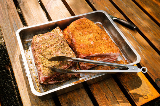 St. Louis Cut Ribs (fleischige Bauchrippen vom Schwein), halbiert, gew&uuml;rzt in Edelstahlbr&auml;ter mit Grillzange auf Holztisch