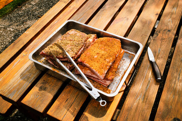 St. Louis Cut Ribs (fleischige Bauchrippen vom Schwein), halbiert, gewürzt in Edelstahlbräter mit Grillzange auf Holztisch