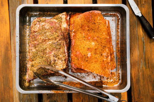 St. Louis Cut Ribs (fleischige Bauchrippen vom Schwein), halbiert, gew&uuml;rzt in Edelstahlbr&auml;ter mit Grillzange auf Holztisch