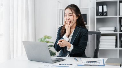 Tired Asian Businesswoman Yawns At Desk Working Overtime on Laptop in Office. Exhausted Adult Female Worker Experiences Fatigue and Needs Rest or Break From Work Overload
