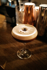 Espresso Martini (Cocktail) mit 3 Kaffeebohnen (bringen Gl&uuml;ck) auf Holzbar, dahinter Cocktailshaker (unscharf)