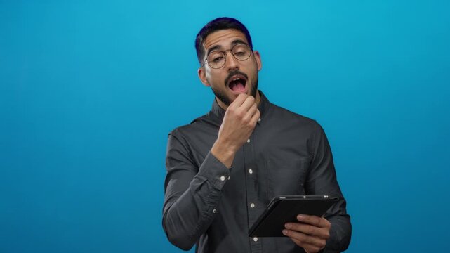 Young man with glasses ponders while holding a tablet against an isolated blue background, displaying a thoughtful expression indicative of having an idea.
