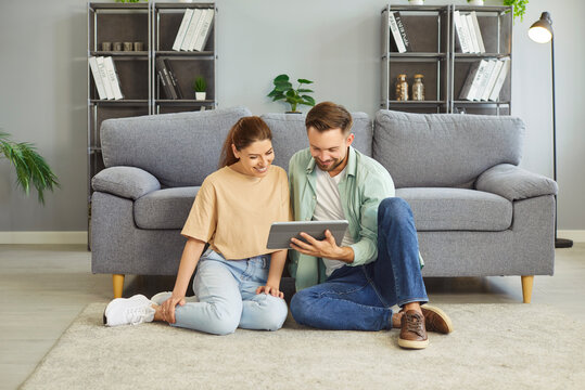 Happy couple using tablet, sitting on living room floor together at home. Young cheerful woman and man watching video in social media or movie on screen, people smiling. Love, bonding concept