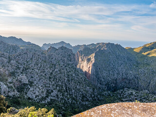 Serra de Tramuntana, Mallorca breathtaking mountain landscape with rugged cliffs and a deep valley bathed in sunlight, surrounded by rolling hills and distant mountains under a clear sky, Majorca