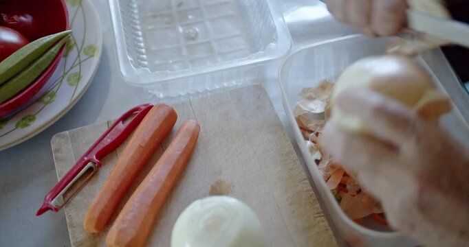 An older person's hands carefully peel a fresh onion on a cutting board, preparing ingredients for a healthy meal. Peeled carrots and okra are also visible, highlighting the process of cooking.