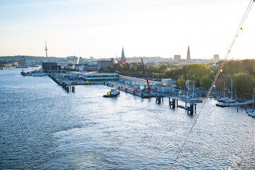 Kiel, serene harbor scene with boats docked, cranes, and a city skyline in the background, with a tower and spires visible in the distance, Germany
