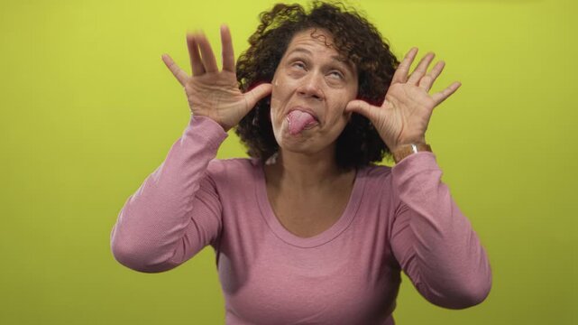 Middle aged woman pulls her hands to her ears while sticking out her tongue in a green studio setting; playfulness.